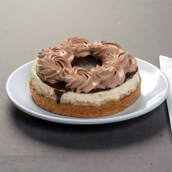 A round cheesecake with a graham cracker crust, topped with chocolate ganache and chocolate whipped cream.