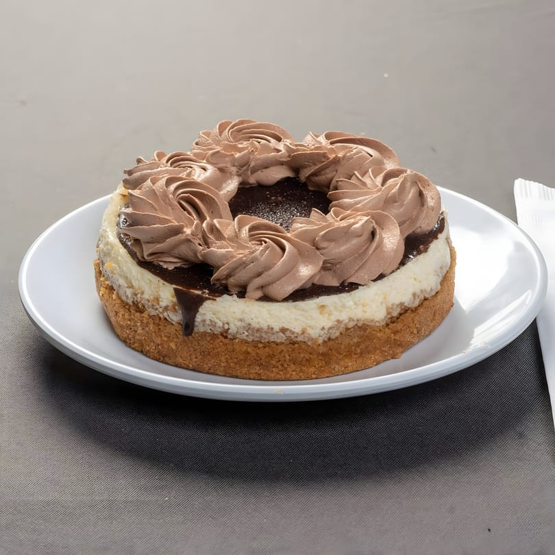 A round cheesecake with a graham cracker crust, topped with chocolate ganache and chocolate whipped cream.
