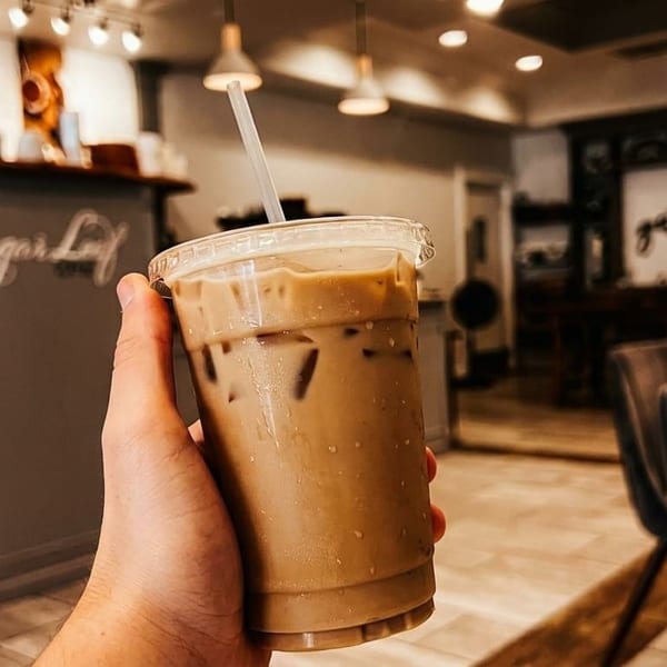 Hand holding an iced coffee with a straw inside a coffee shop with a "Sugar Loaf" counter.